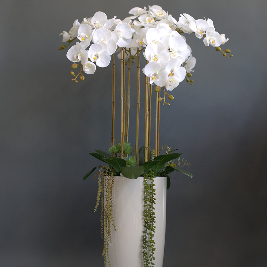 7 белых, искусственных фаленопсиса в высоком, напольном кашпо.